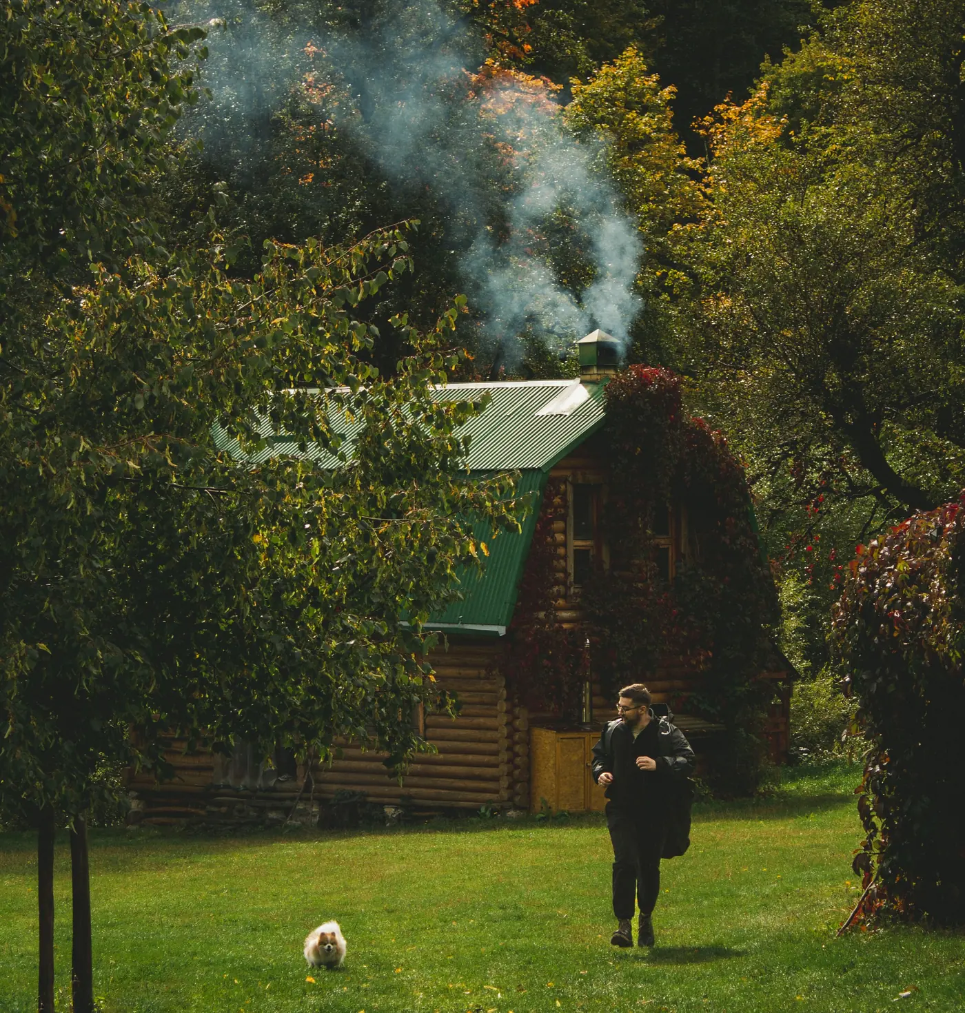 Woodsmoke rising from the chimney of a log cabin in autumn woodland, a man and his dog crossing the grass outside