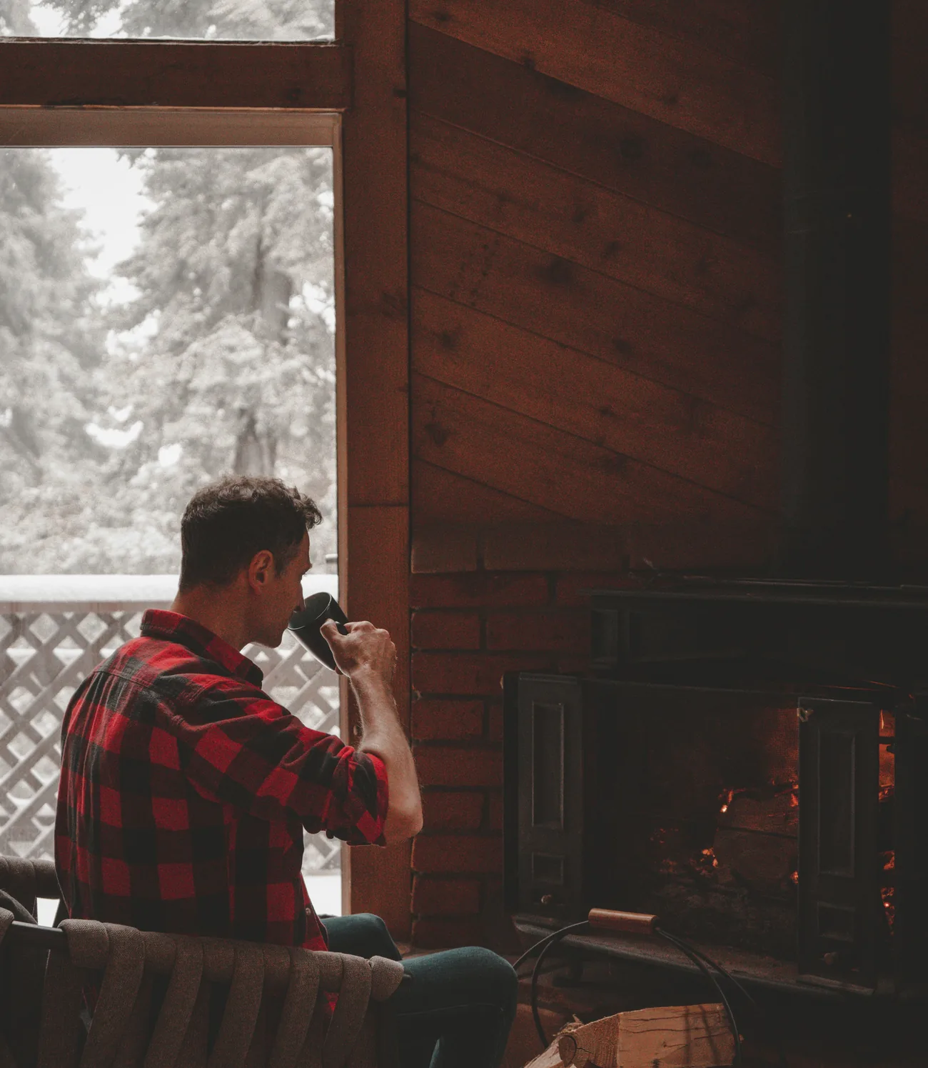 A lodge owner sitting with a mug beside a wood stove, snow through the window